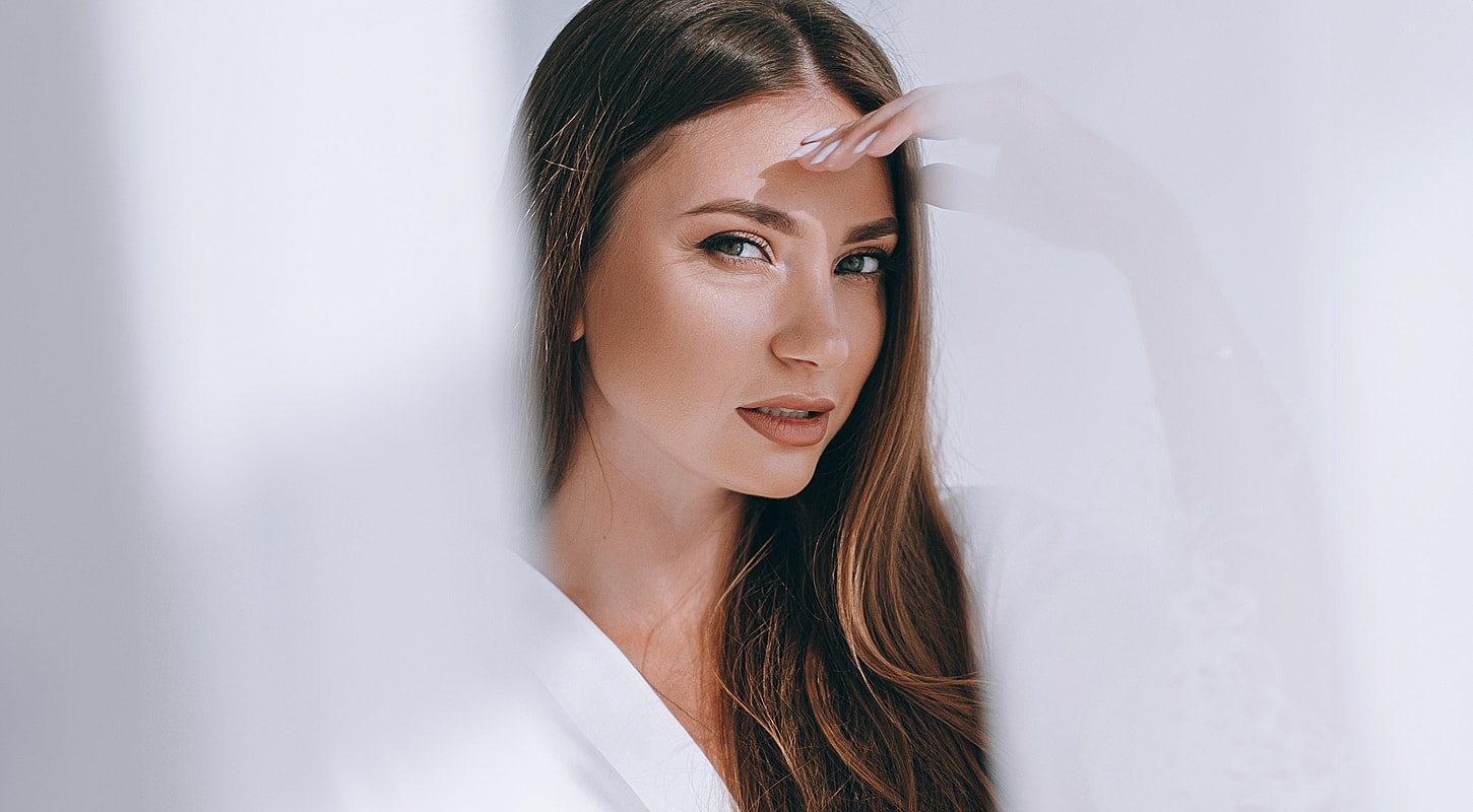 Woman with long hair peeking through sheer curtains.